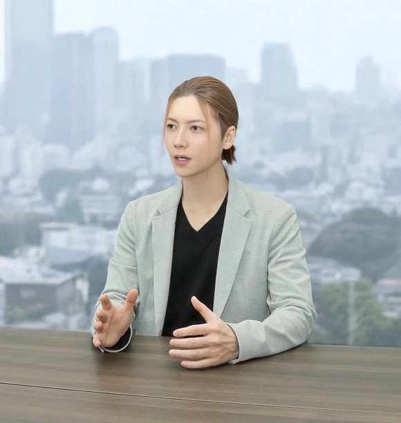 窓を背に、テーブルに座って話す男性。